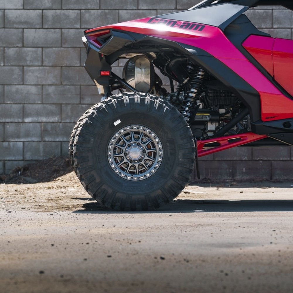 A close-up side view of a pink and black Can-Am off-road vehicle, focusing on the rear Fuel UTV Cycle utv wheel in gloss gunmetal color set against a gray concrete block wall.