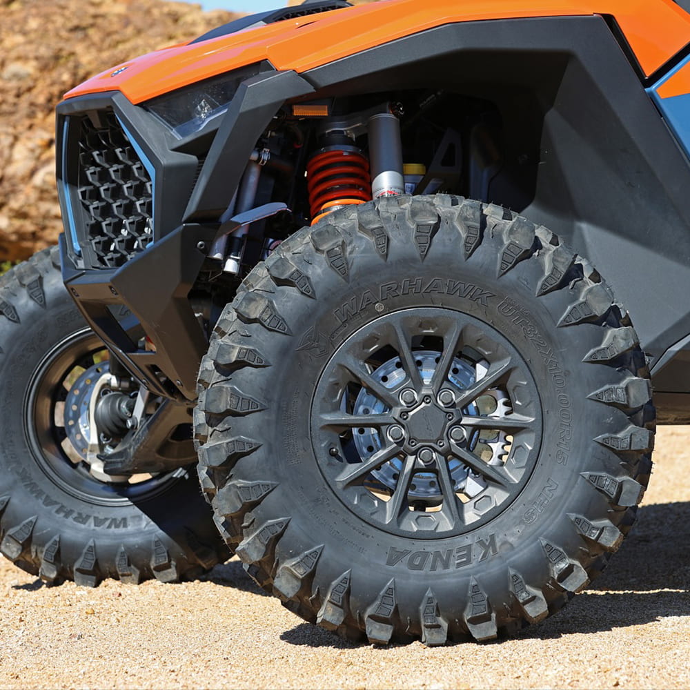 Sidewall view of the Kenda Warhawk UTV tires mounted and installed on an orange Polaris RZR, parked on sand and gravel, ready to tackle any terrain, showcasing the stepped down shoulder lug design for technical off road riding.
