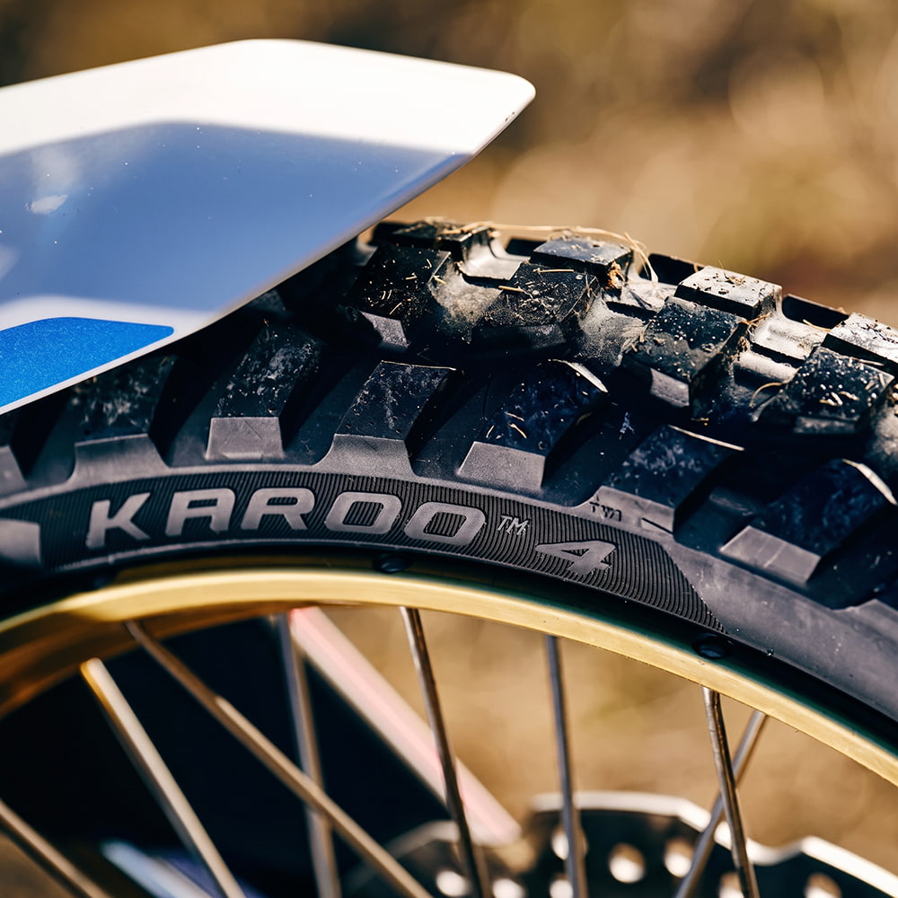 Close-up of a Metzeler Karoo 4 dual sport tire mounted on a motorcycle wheel, with a white and blue fender visible above and a blurred outdoor background.