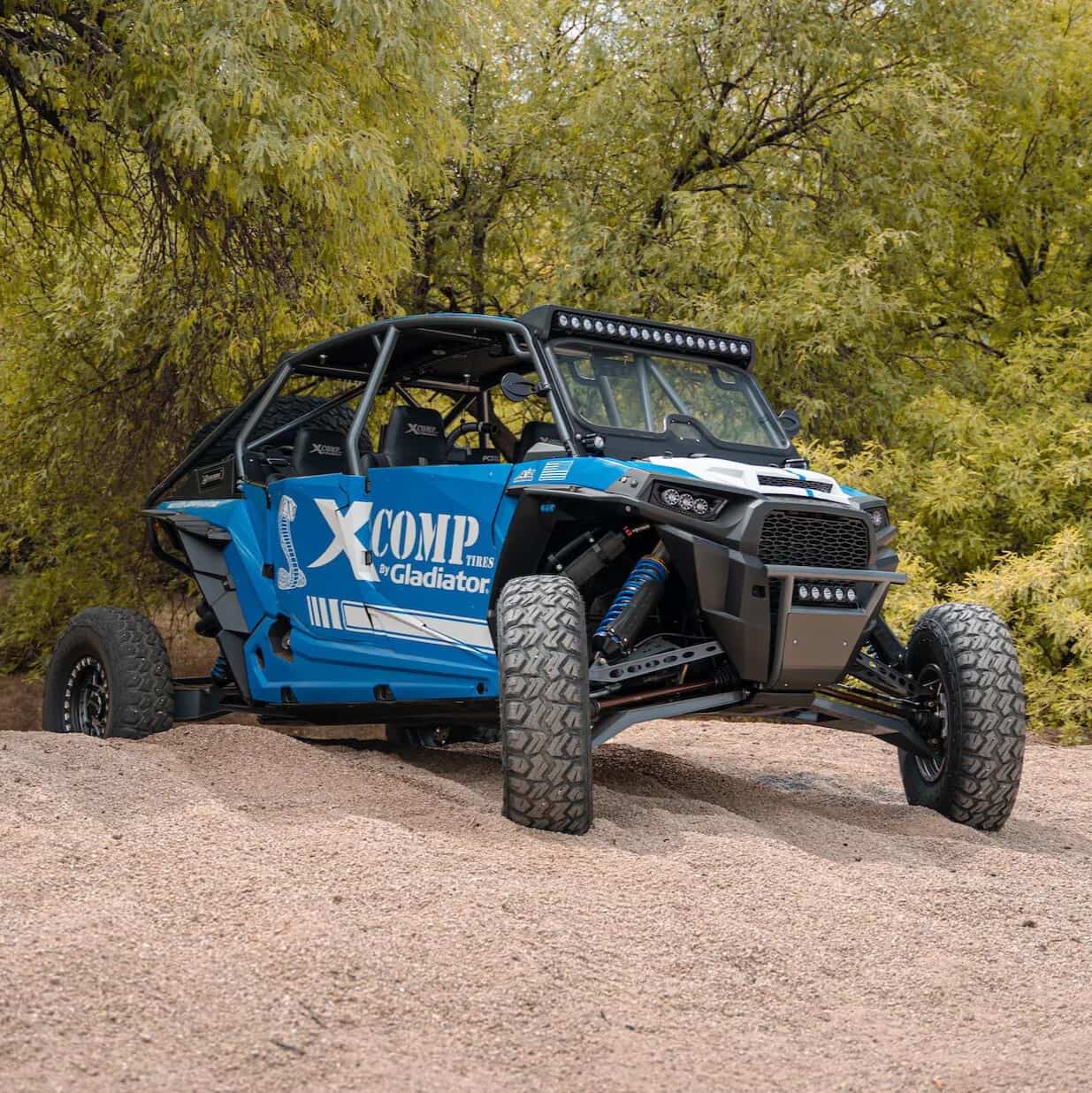 XComp R/S 10-Ply SBR high performance all-terrain tires mounted on Polaris RZR SXS with beadlock 15" rims, parked on sand bar with trees in the background.