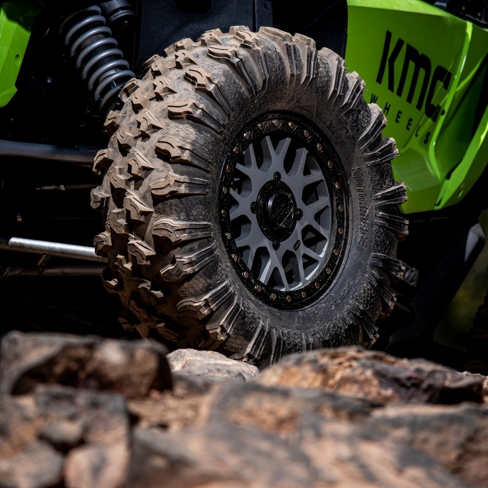 Close-up of the EFX MotoRavage off-road UTV tire with deep treads mounted on a silver multi-spoke wheel, attached to a bright green KMC Wheels vehicle on rocky terrain.
