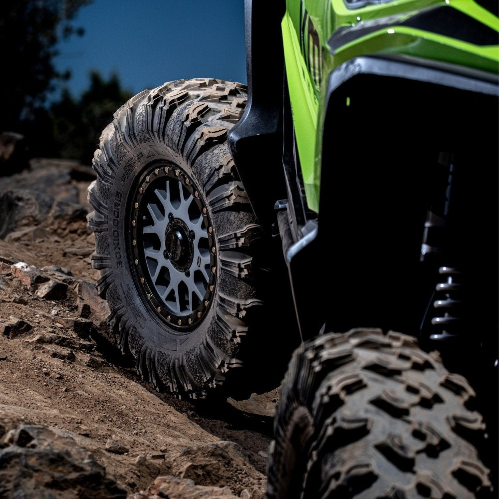 Close-up of the EFX MotoRavage UTV tire with aggressive tread and a silver multi-spoke wheel, attached to a bright green and black all-terrain vehicle on a rocky dirt trail.