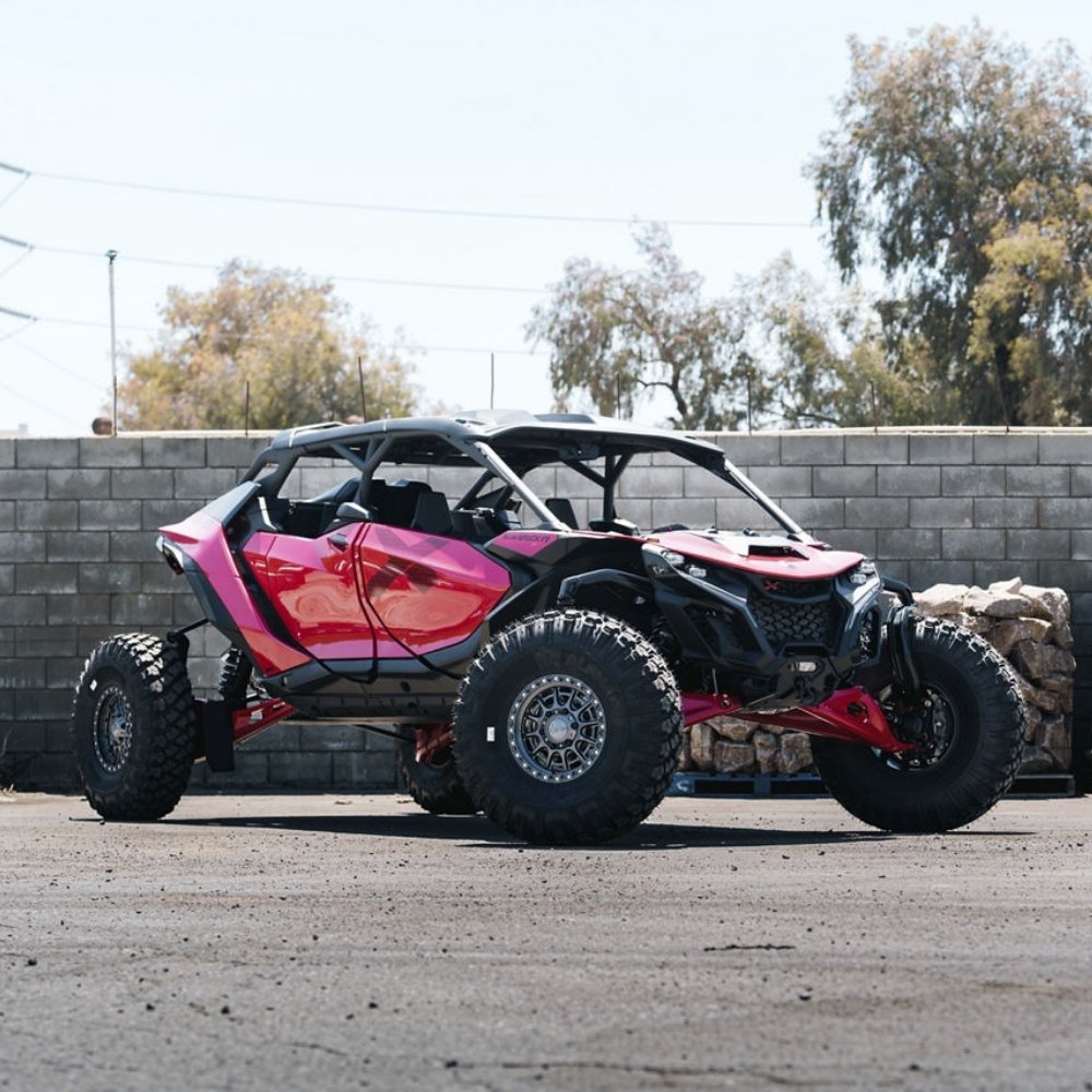 A pink and black off-road side-by-side UTV with large knobby tires and Fuel UTV Cycle utv wheels in gloss gunmetal color, parked outdoors on a paved surface in front of a cinderblock wall and trees.
