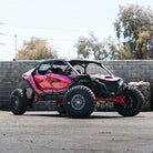 A pink and black off-road side-by-side UTV with large knobby tires and Fuel UTV Cycle utv wheels in gloss gunmetal color, parked outdoors on a paved surface in front of a cinderblock wall and trees.