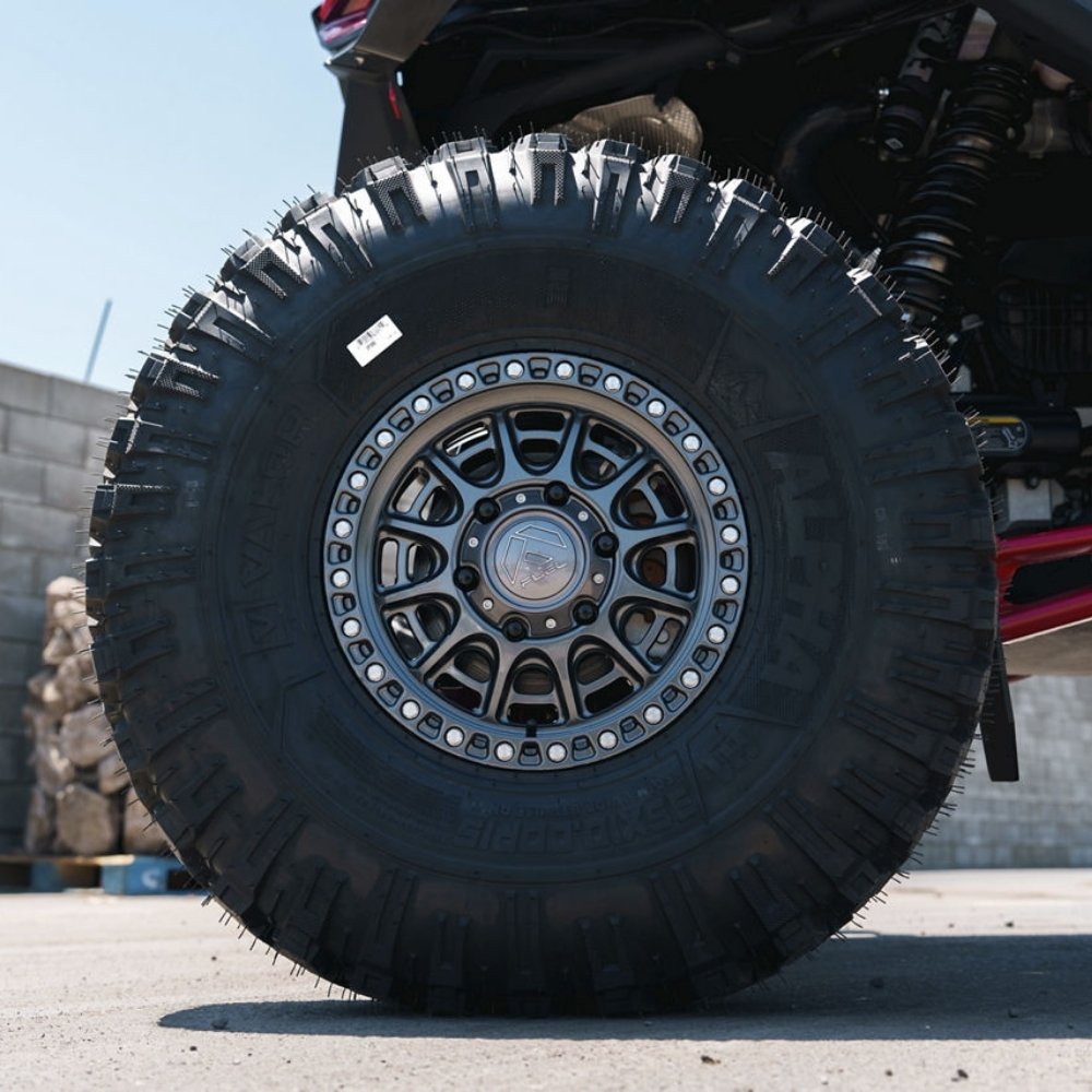 A close-up shot of a black off-road tire mounted on a Fuel UTV Cycle utv wheel in gloss gunmetal attached to a UTV undercarriage, photographed outdoors on a paved surface.