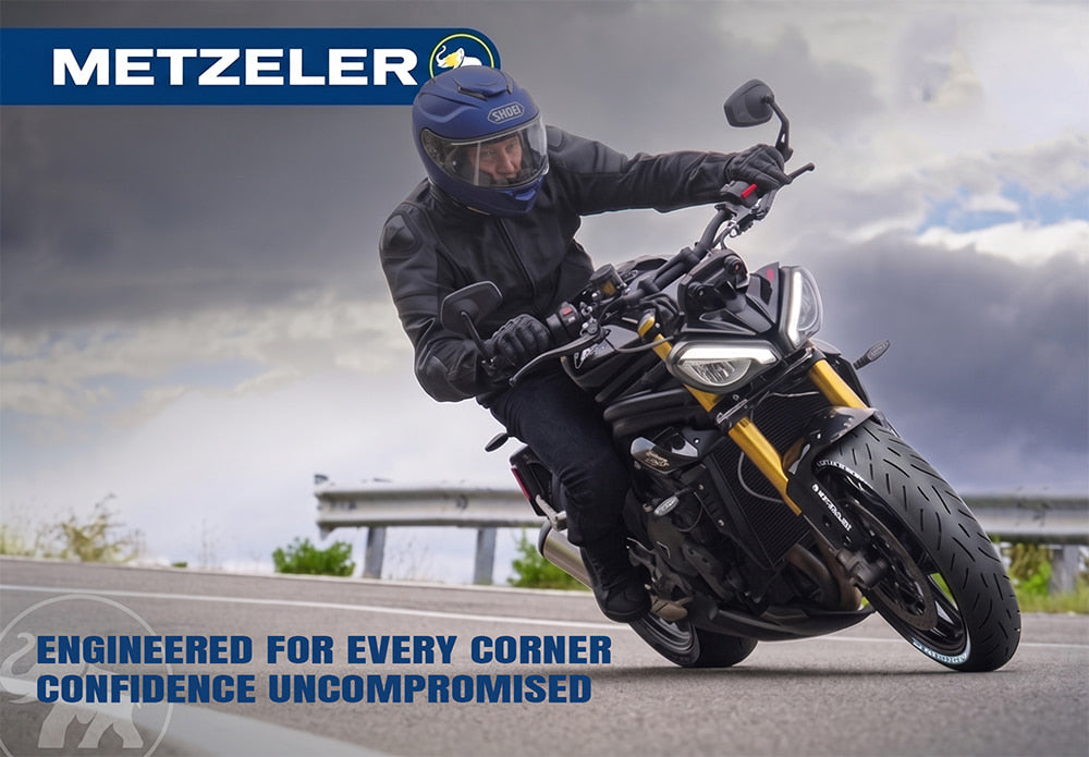 A motorcycle rider in black gear and a blue helmet leans into a turn on a black and gold motorcycle on an open road, with the Metzeler logo and slogan "Engineered for every corner. Confidence uncompromised".