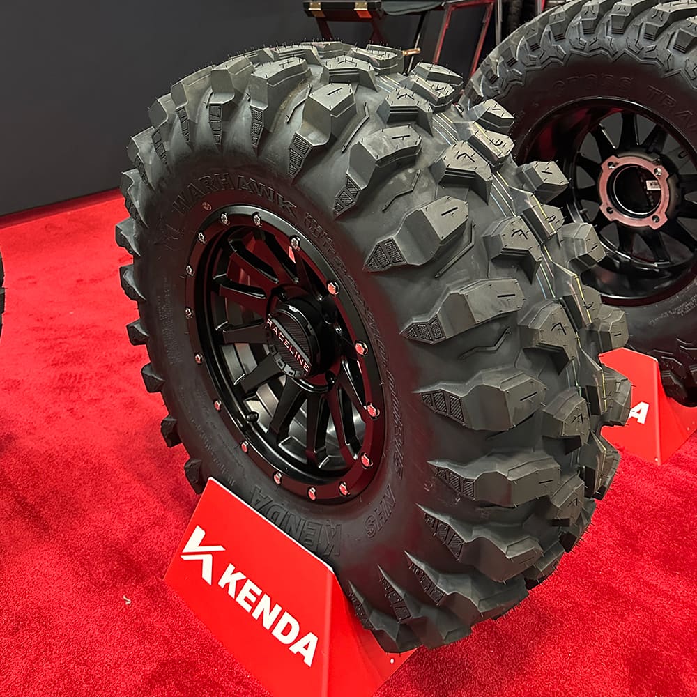 Brand new UTV tire, the Kenda Warhawk 8-ply nylon radial heavy duty aggressive off road and all terrain tire, featuring two new sizes, 32x10R14 and 33x10R15, shown here mounted on a Raceline wheel, displayed at a trade show in the Kenda booth, on a red carpet, sold by Ride or Die Tire.
