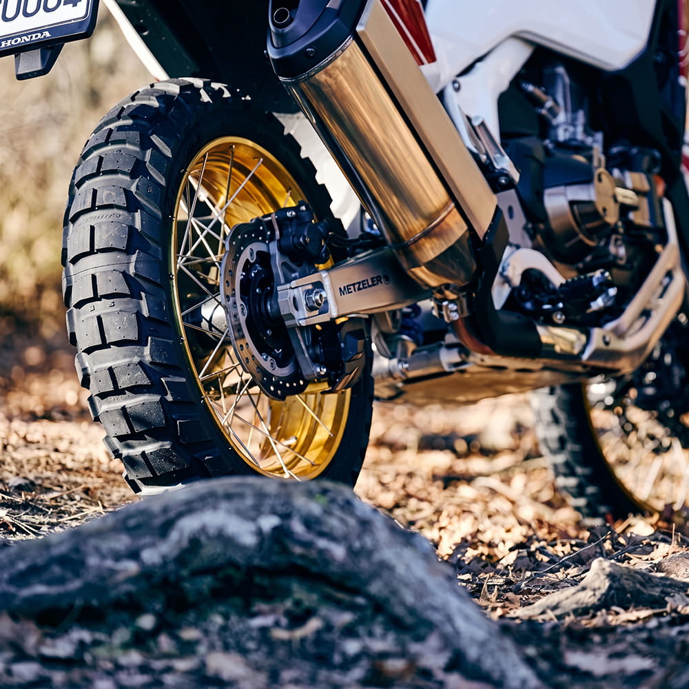 Close-up of a motorcycle's rear Metzeler Karoo 4 tire and exhaust system outdoors on a dirt road. The wheel has a gold rim, the tire is a dual sport adv design.