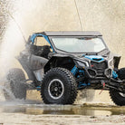 A black and blue Polaris RZR SXS with SuperATV Incursion AT 32x10R15 tires driving through a creek, creating a spray of mist.