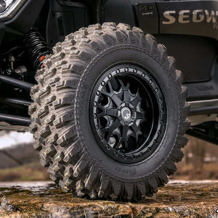 A close up view of a SuperATV XT Warrior tire mounted on a SuperATV beadlock wheel on a Segway SX10 SXS, parked on a rocky wet ledge.
